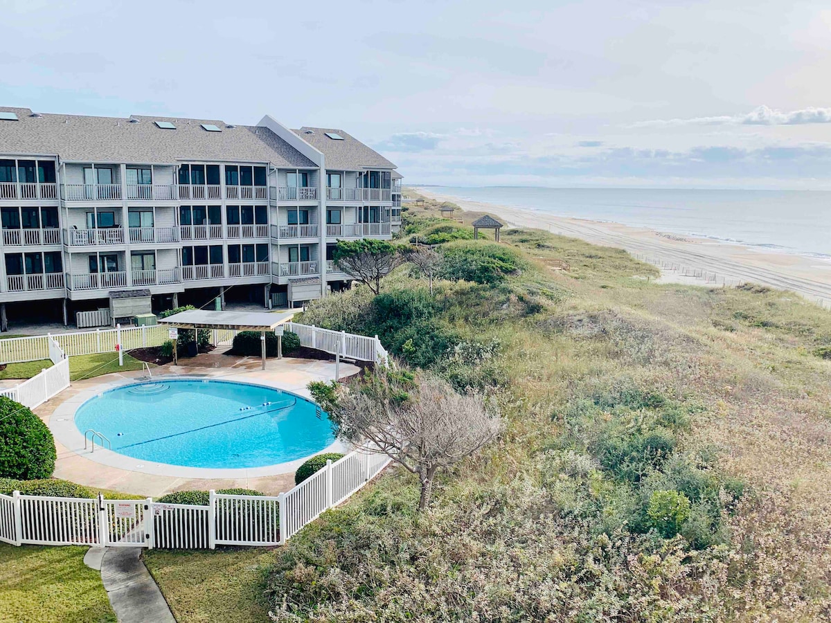 A view from above captures a circular swimming pool surrounded by a white fence and lush landscaping, with the beach visible in the distance. The modern three-story building features balconies, providing a backdrop to the tranquil scene of sand and ocean waves.