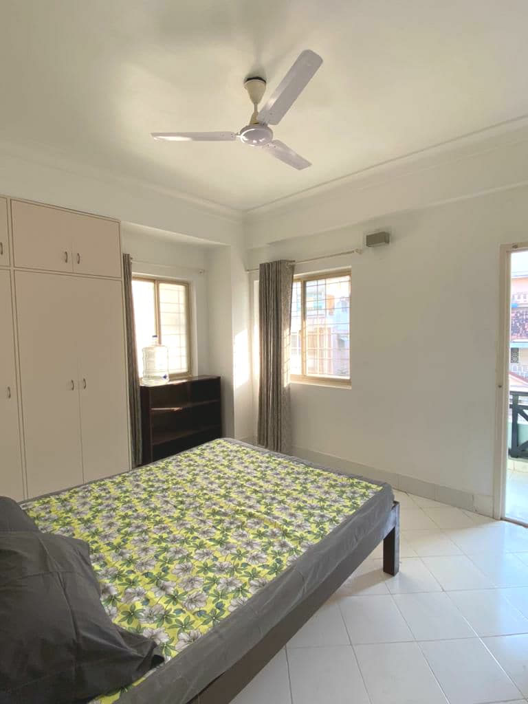 The bedroom features a spacious bed adorned with a patterned bedspread. Natural light enters through two windows, enhancing the bright ambiance. A ceiling fan is positioned above, and a light-colored wardrobe and a dark shelf are visible against the wall.