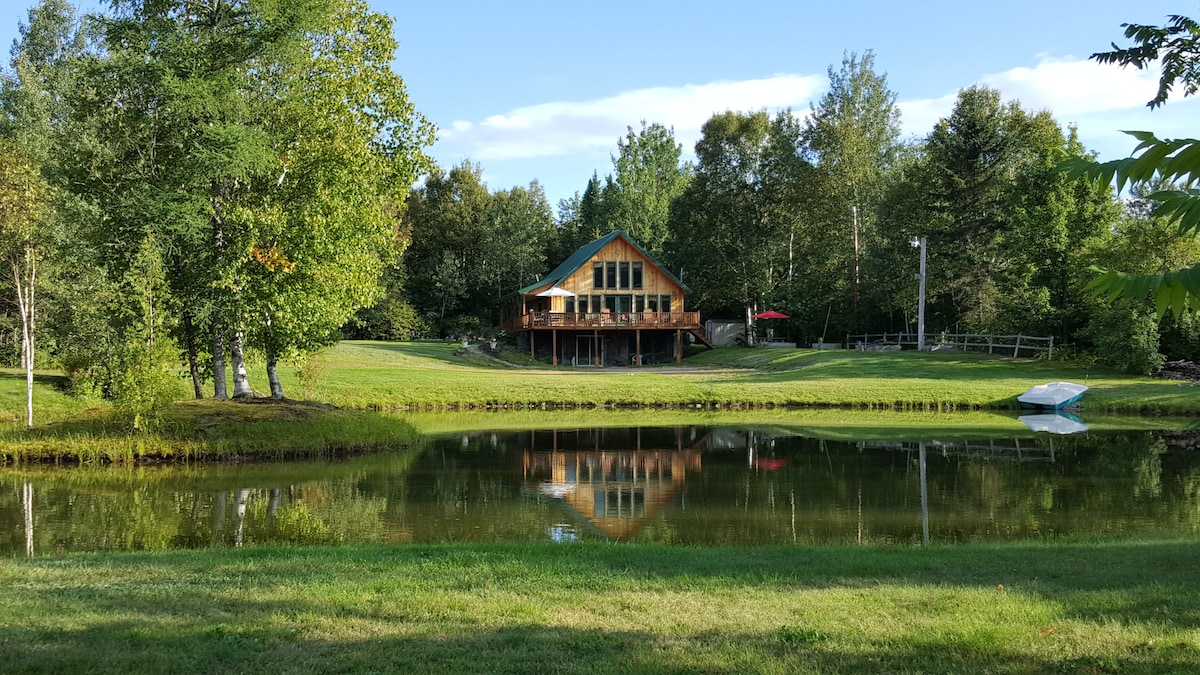 A charming chalet is reflected on the tranquil surface of a private trout pond. Surrounding greenery includes trees and manicured lawns. The building features a sloped roof and large windows, allowing natural light to enter. A small boat is visible near the water's edge.
