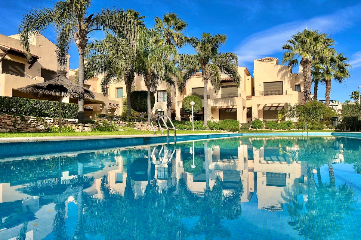 A serene communal pool reflects the surrounding palm trees and architecture, under a clear blue sky. Sun loungers are positioned beside the water, while lush greenery complements the tranquil atmosphere. This outdoor space offers a refreshing escape for relaxation.