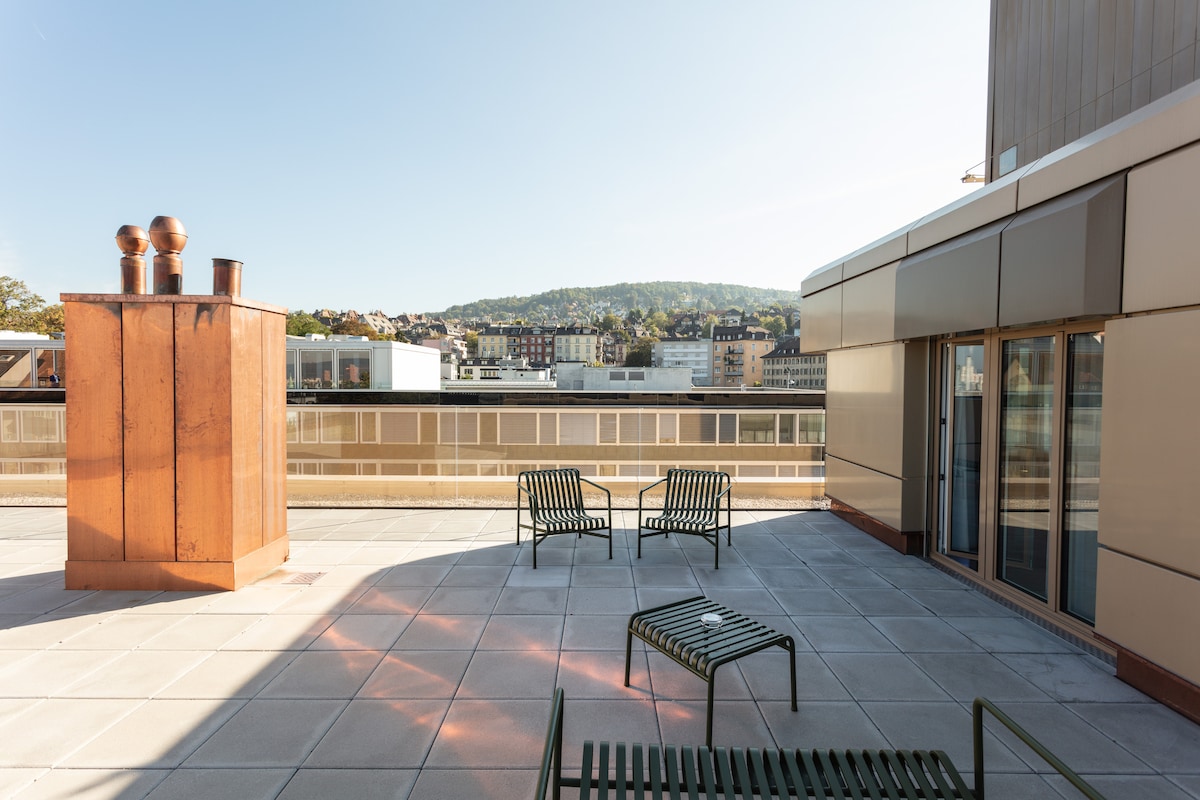 A spacious rooftop terrace features two metal lounge chairs, offering views of the surrounding area, including a hillside. Sunlight casts shadows on the light-colored paving stones, while a copper feature provides a distinctive touch to the outdoor space.