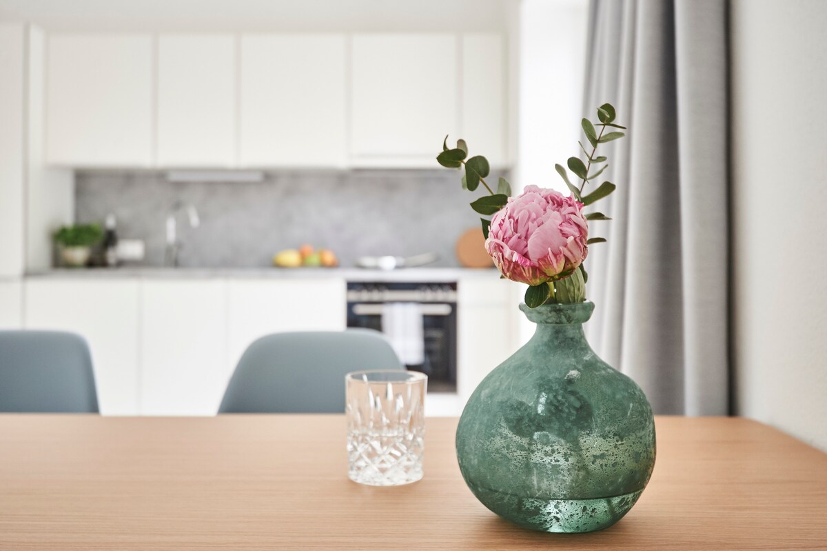A dining table with a wooden surface features a green glass vase holding a pink flower. A clear glass tumbler rests beside the vase. In the background, a well-equipped kitchen is visible, showcasing modern appliances and a subtle color palette.