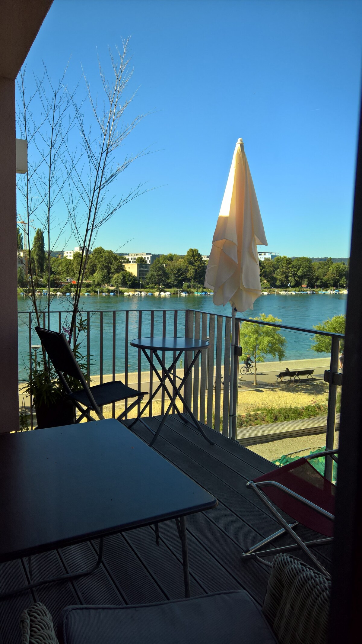 A balcony is visible, featuring a small table and chairs. A large umbrella provides shade. The serene view includes the calm waters of the Rhine, lined with trees and buildings in the background. Clear blue skies enhance the outdoor ambiance.