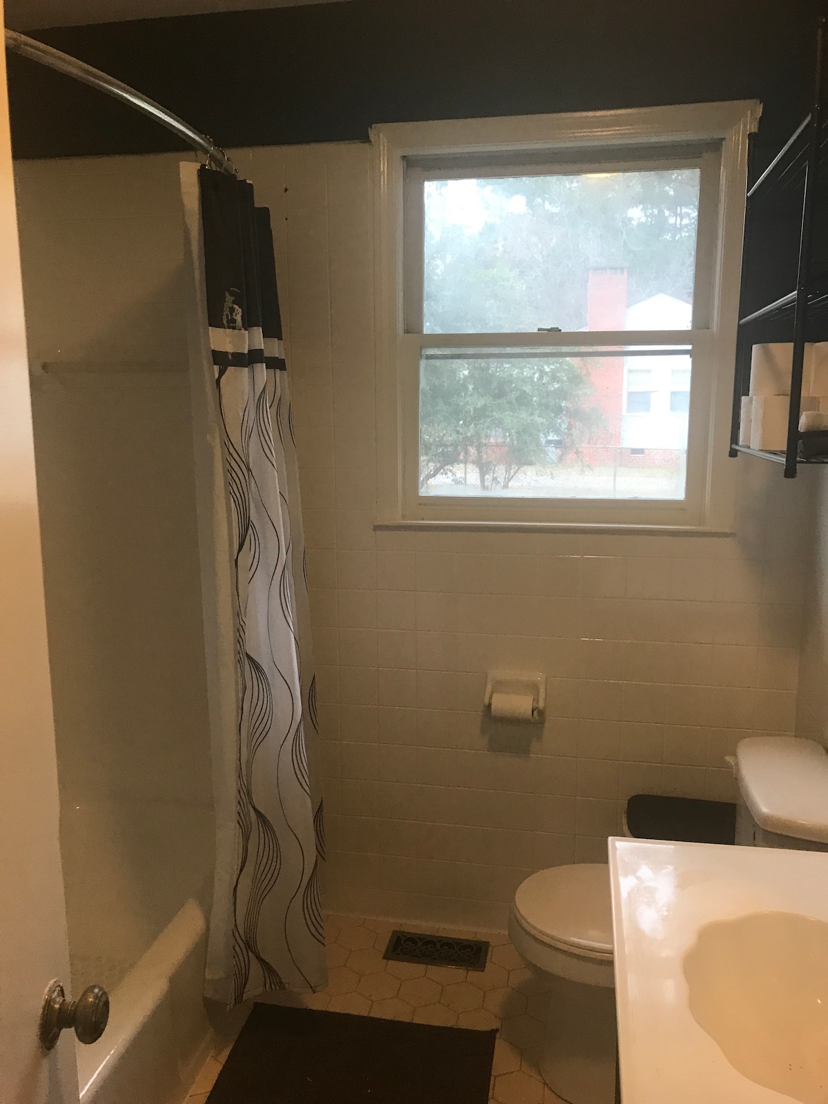 The bathroom features a combination tub and shower with a patterned curtain, a single sink with a countertop, and a toilet. A window allows natural light to enter, offering a view of the outside. Basic toiletries and a bath mat are visible.