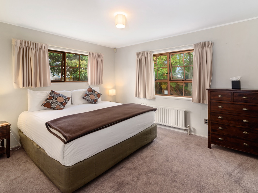 A well-appointed bedroom features a spacious bed with decorative pillows, complemented by warm lighting from bedside lamps. Two windows allow natural light in, framed by soft drapes. A wooden dresser adds storage, with a neutral carpet covering the floor.