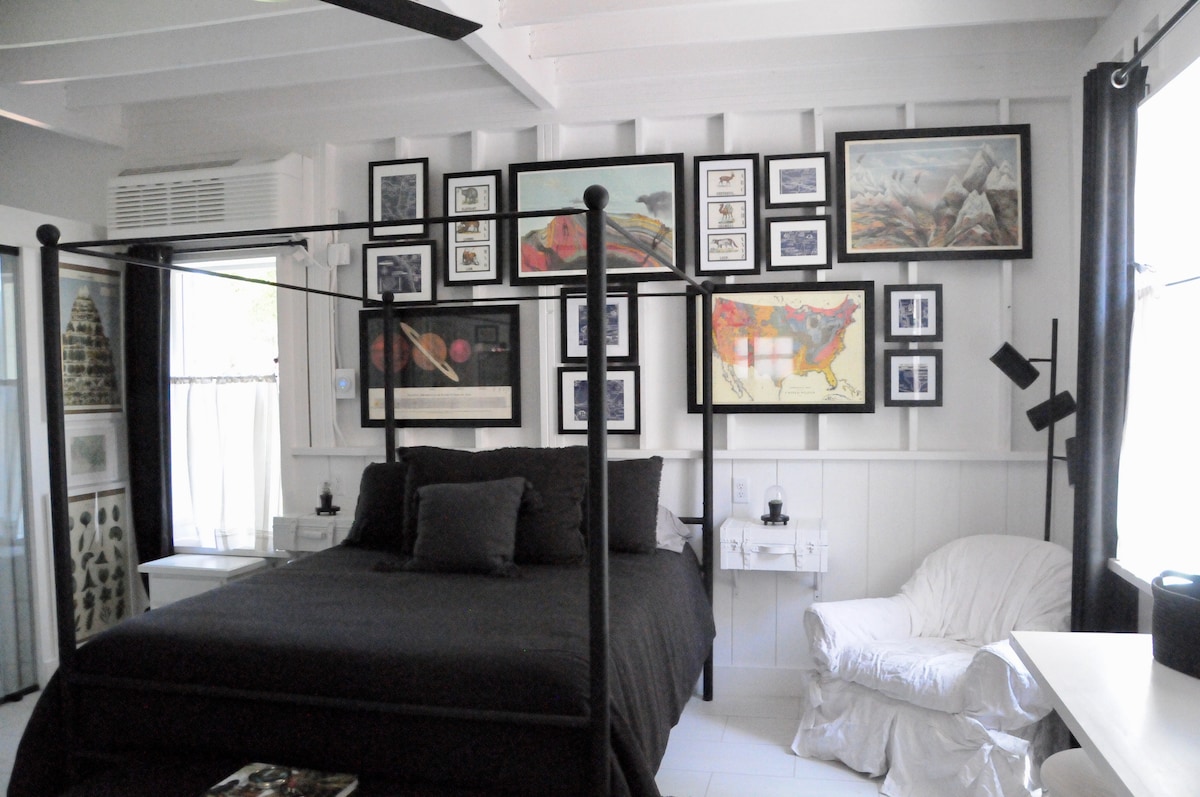 A black canopy bed occupies the center of the room, flanked by stylish white nightstands. The wall is adorned with an eclectic collection of framed artwork and maps. A white armchair is positioned in one corner, complemented by natural light from nearby windows.