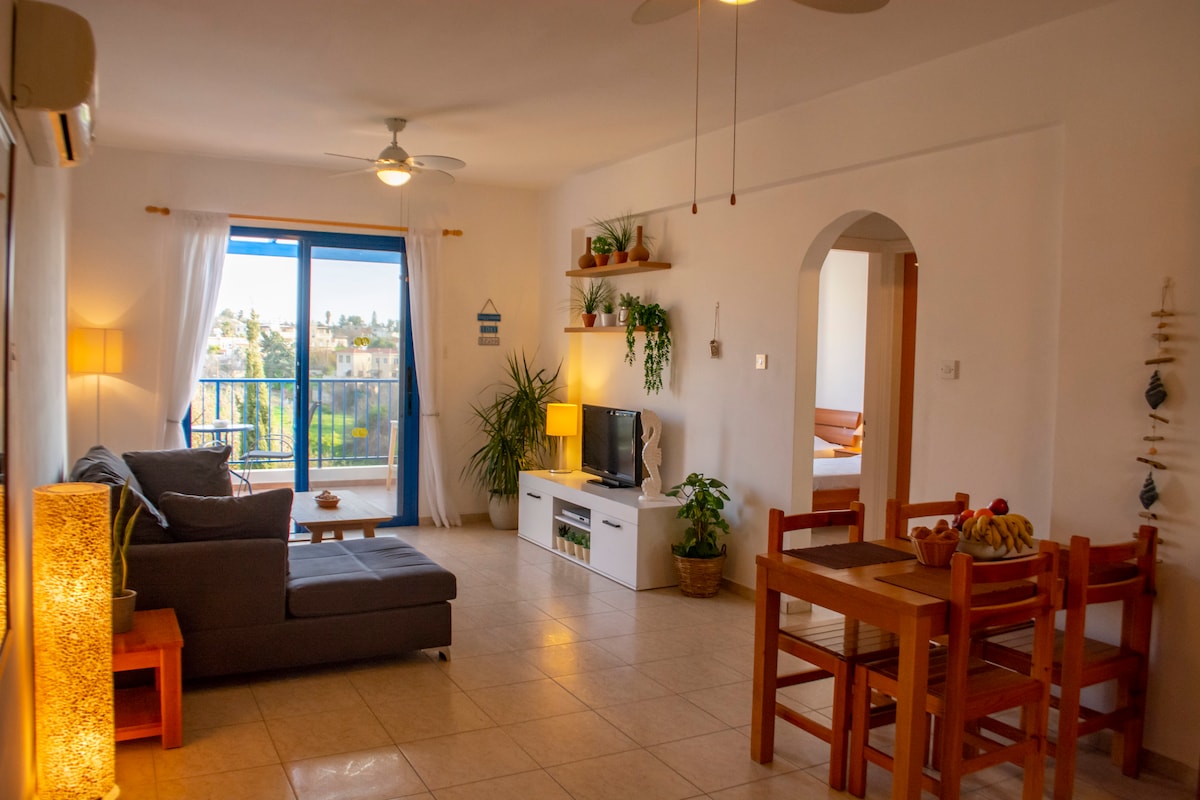 A bright living area features a comfortable gray sofa and a wooden dining table surrounded by four chairs. A television is positioned against the wall, complemented by potted plants. Large windows offer a view of the greenery outside, with natural light illuminating the space.