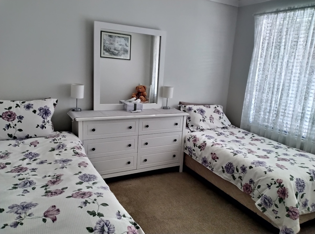 A serene sleeping area features two beds adorned with floral-patterned bedding. A white dresser with six drawers sits against the wall, accompanied by two bedside lamps. A large mirror reflects the soft natural light coming through a window dressed with sheer curtains.