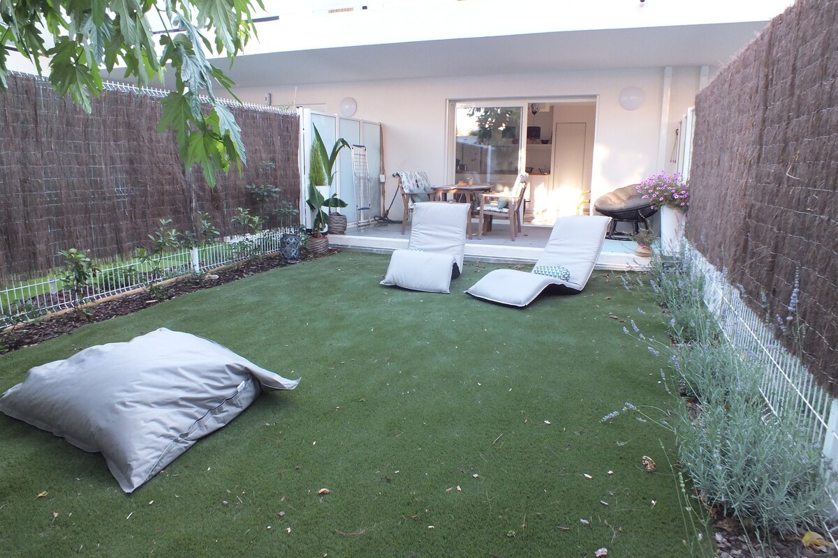 The garden area features a lush green lawn with two reclining lounge chairs positioned for relaxation. A small table and additional seating are visible, complemented by potted plants. The space is enclosed by natural fencing, creating a tranquil and private atmosphere.