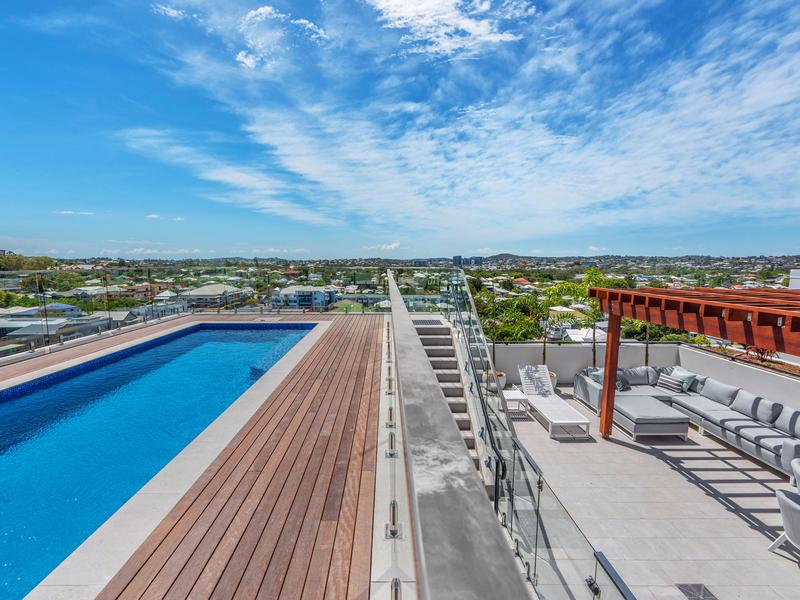 The rooftop area features a large swimming pool adjacent to a spacious deck. Surrounding views of the city and landscape are visible under a clear blue sky. Comfortable seating is provided in a shaded area, designed for relaxation and social gatherings.