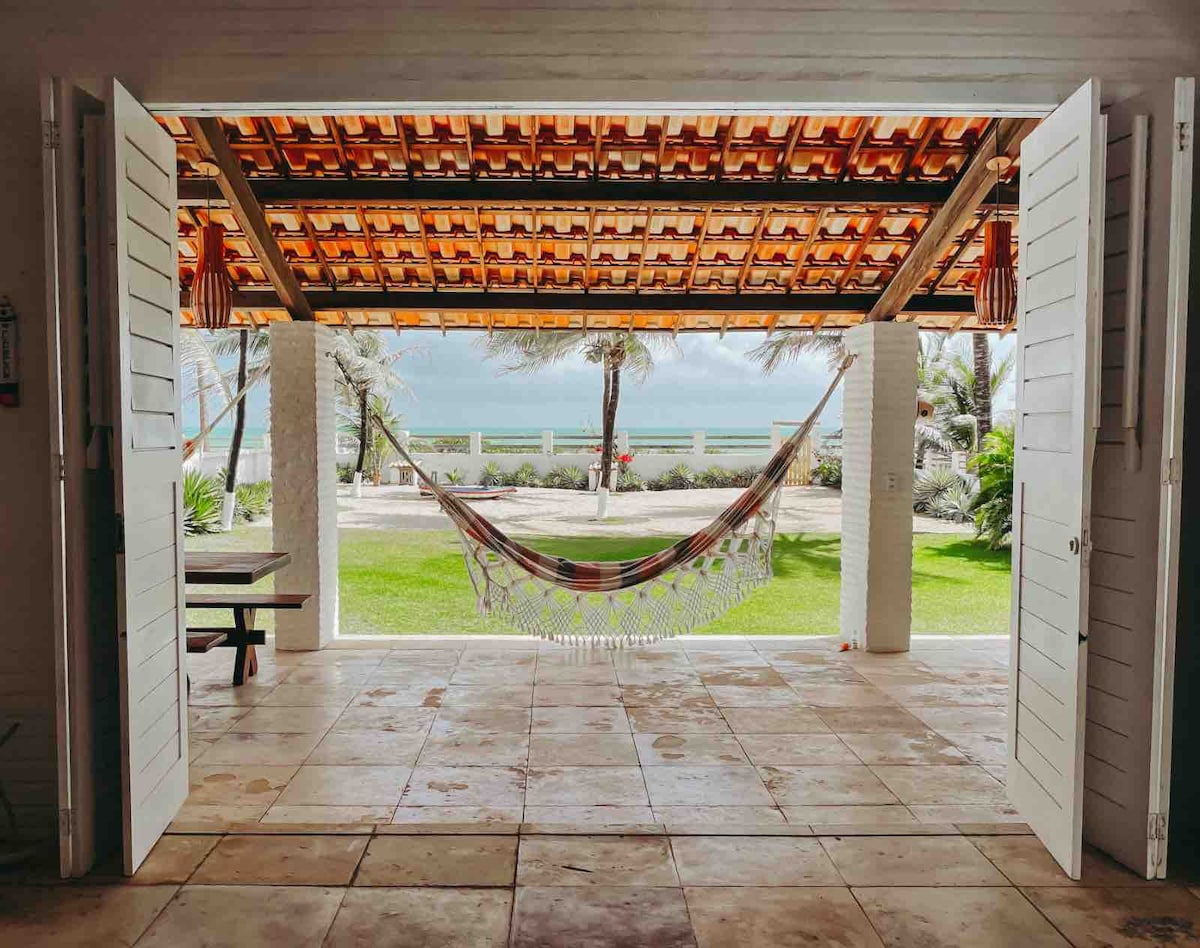 A spacious terrace area is framed by open double doors, revealing a lush green garden and the beach view beyond. A colorful hammock is hung under a thatched roof, inviting relaxation. The tiled floor reflects natural light, enhancing the serene ambiance of the space.