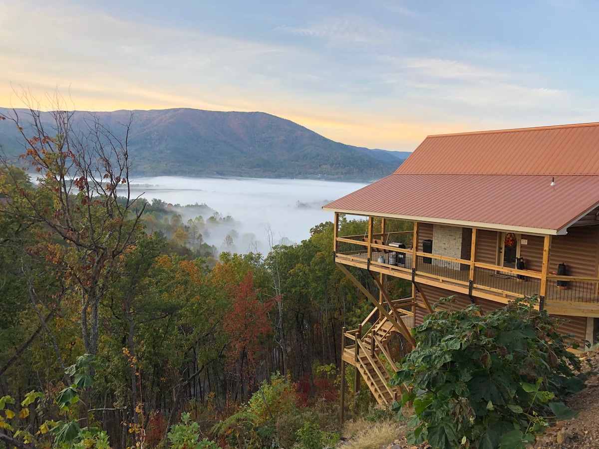 A cabin is positioned on a hillside, overlooking a misty valley. Lush trees surround the structure, while colorful foliage hints at the changing seasons. The elevated deck invites relaxation, providing vantage points for enjoying the surrounding natural beauty and expansive mountain views.
