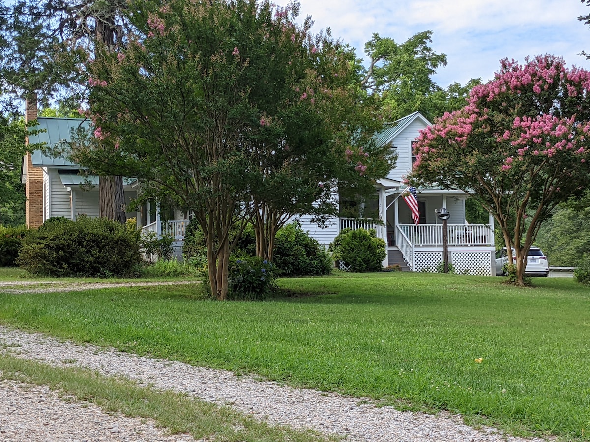A country home is set amidst manicured lawns and flowering trees, featuring a welcoming porch with an American flag. The gravel path leads to the entrance, and the property is surrounded by greenery, providing a serene rural atmosphere.