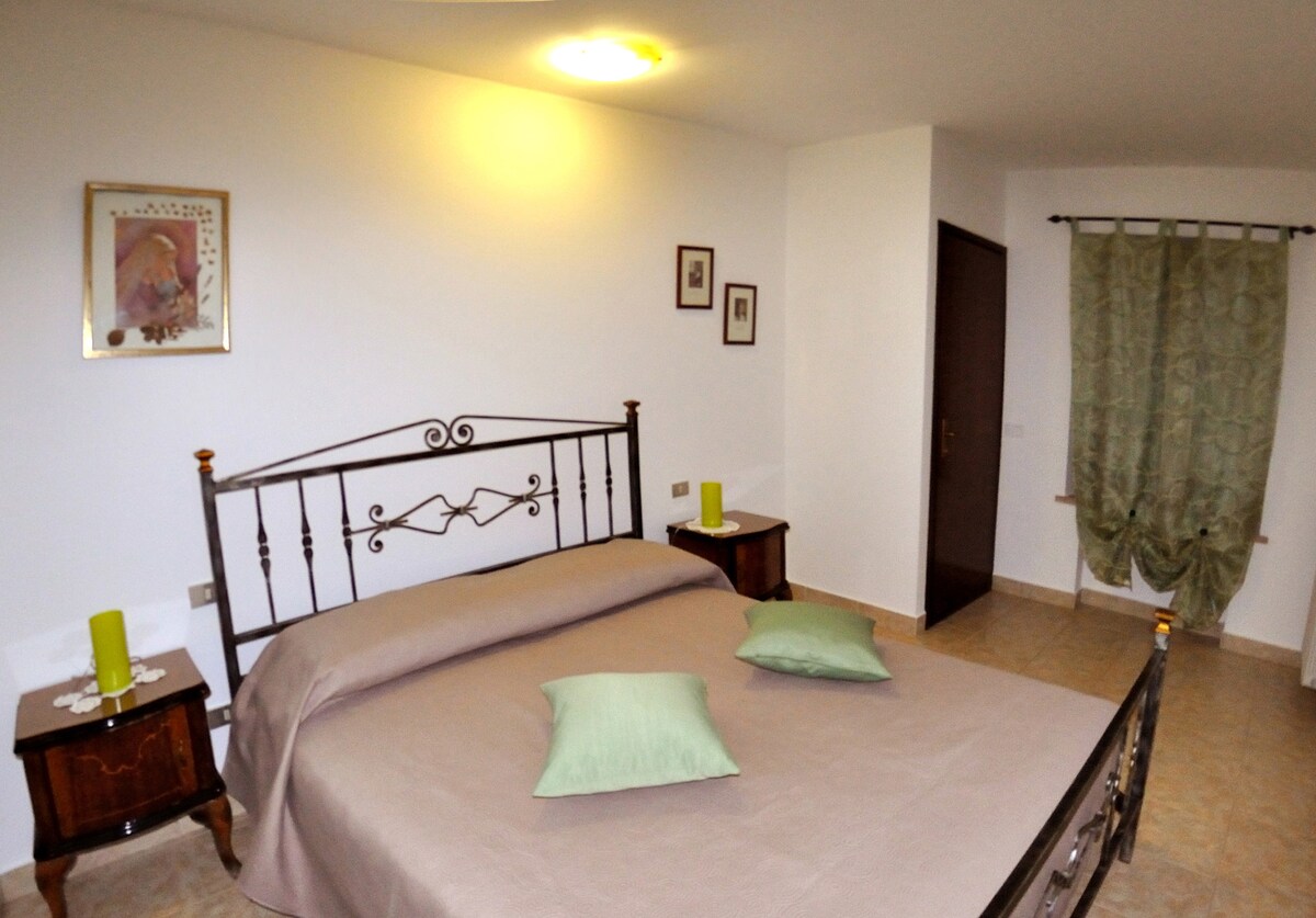 A spacious sleeping area features a metal bed frame with a neutral-colored blanket. Two green decorative pillows are placed on the bed. Dark wooden side tables are positioned on either side, complemented by green lamps. Soft lighting enhances the calm ambiance.