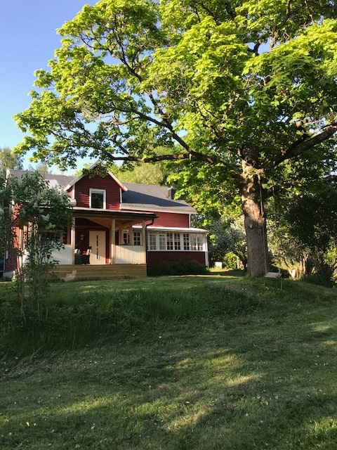 A traditional two-story red house is framed by a large green tree and lush lawn. The porch is visible, providing a welcoming entryway, while the surrounding foliage adds a natural backdrop, creating a serene outdoor environment.