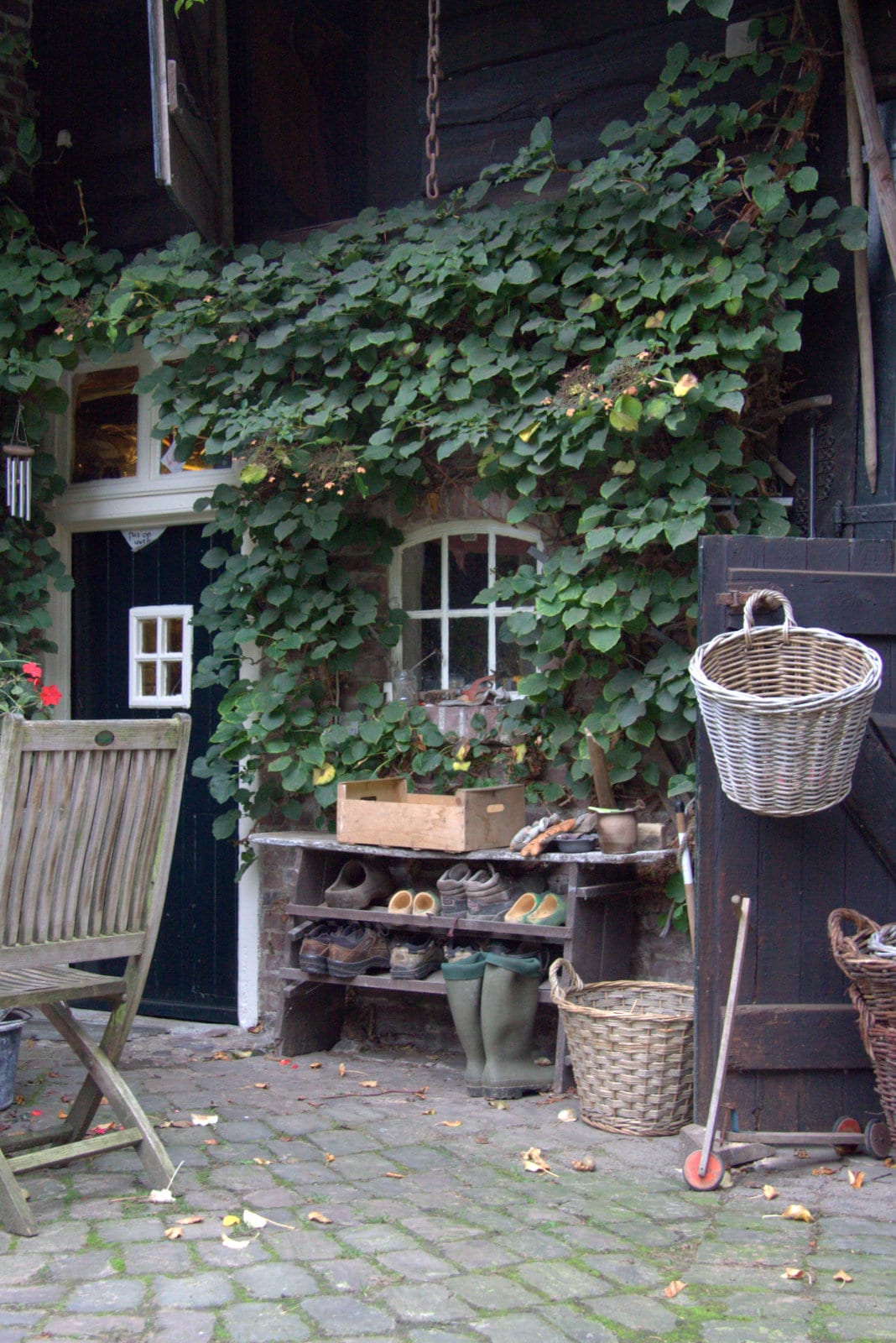 The entrance to a charming courtyard is adorned with lush greenery, framing a small window and door. A wooden shelf displays gardening shoes and tools, complemented by woven baskets. Weathered stone pathways are visible, adding to the rustic atmosphere of the space.