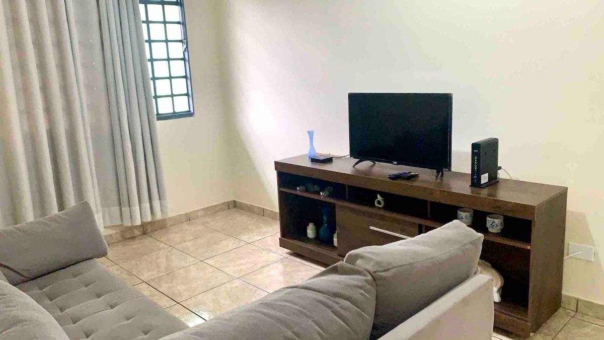 A comfortable living room is featured, showcasing a beige sofa positioned near a low wooden TV stand. A television sits atop the stand, accompanied by decorative items. Light filters through a window covered by sheer curtains, illuminating the tiled floor.
