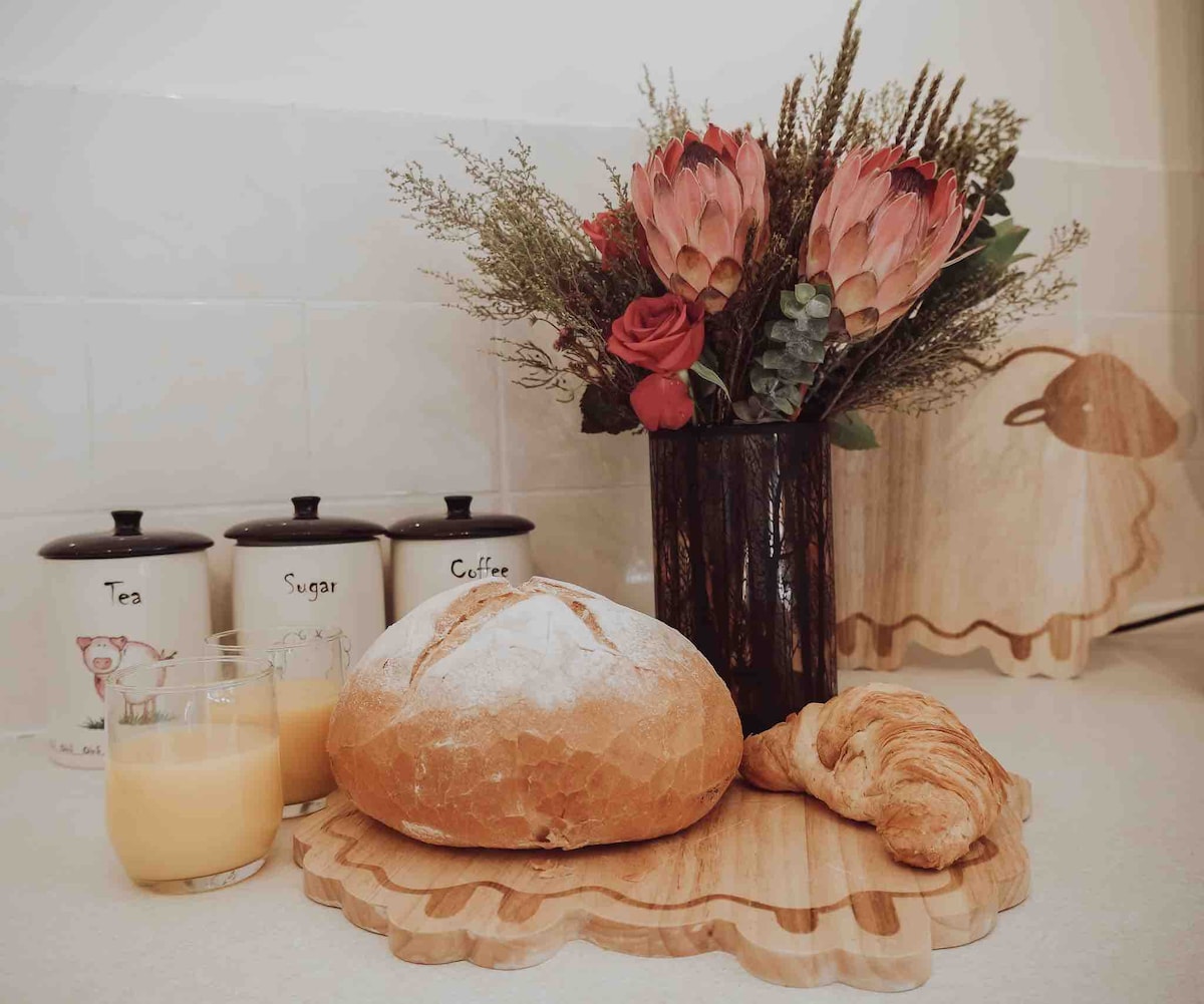 A wooden platter presents a round loaf of bread and a buttery croissant. Two glasses with a light yellow beverage sit nearby. A vase filled with dried flowers and a decorative container labeled 'Tea', 'Sugar', and 'Coffee' add a touch of charm to the scene.