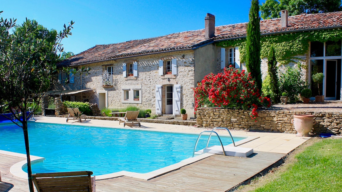 A spacious private pool is framed by a sun terrace, featuring lounge chairs for relaxation. The restored farmhouse showcases a blend of stone and greenery, with blooming flowers and lush foliage enhancing the outdoor space. Sunlight bathes the scene, emphasizing the inviting atmosphere.