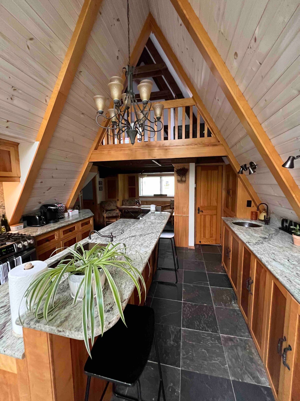 The A-frame kitchen features wooden cabinetry and a spacious granite countertop, accommodating several bar stools. A unique chandelier hangs from the high ceiling, while a window offers natural light. The open layout connects to the dining area and living space, enhancing the overall sense of openness.