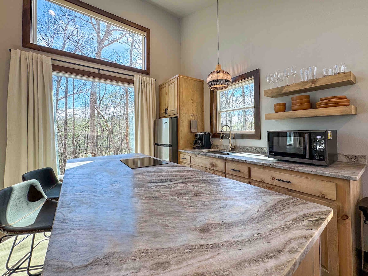 A modern kitchen is displayed, featuring natural wood cabinetry and a spacious granite countertop. Large windows provide ample natural light and views of the surrounding trees. Shelves hold various dishware, while a microwave and coffee maker are positioned on the counter for convenience.