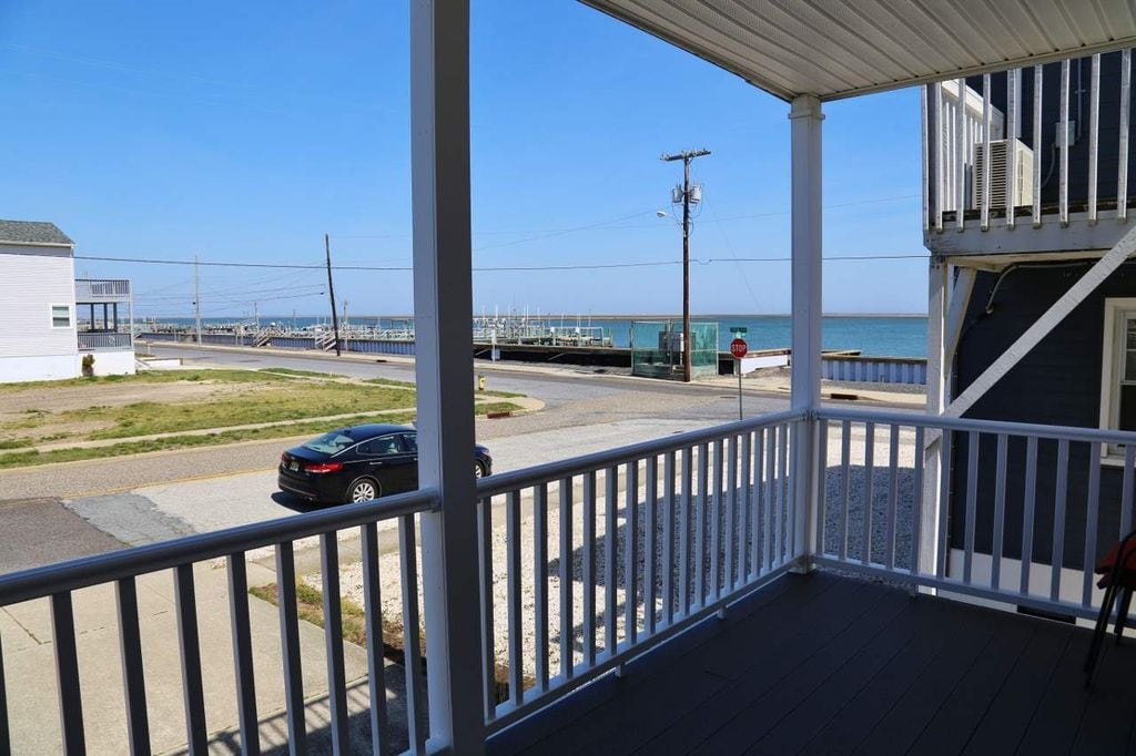 A spacious balcony offers views of the bay and nearby marina. The scene includes a clear blue sky, distant water, and a street with parked vehicles. A white railing frames the perspective, creating an open feel, while soft natural lighting enhances the coastal ambiance.