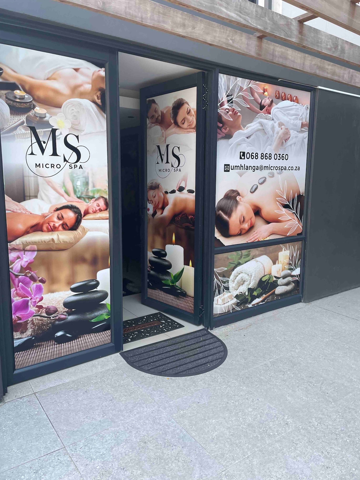 The entrance to a micro spa is depicted, featuring large glass doors adorned with relaxing spa imagery. The design includes various wellness elements such as stones, flowers, and serene massage themes, inviting guests to experience relaxation and rejuvenation.