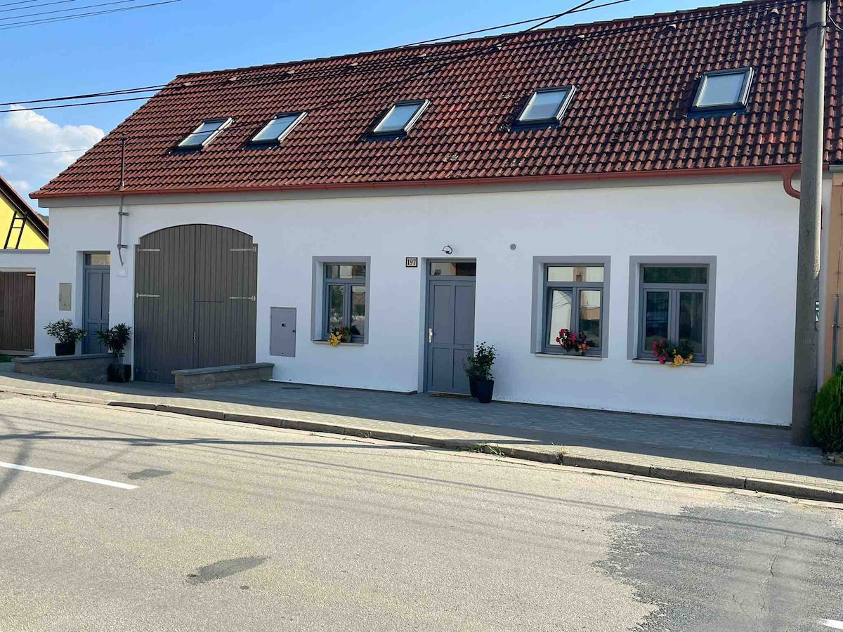 A charming building with a red-tiled roof features multiple windows, allowing natural light to fill the interior. Flower pots are placed under the windows, adding a touch of greenery. An entrance with a wooden door is visible, along with a rustic garage door.