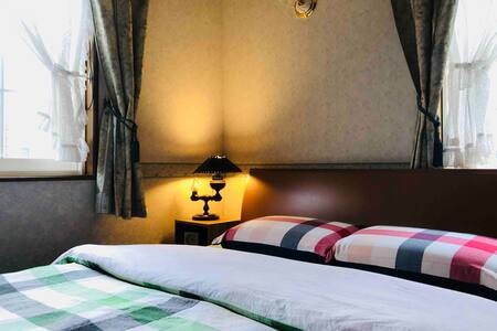 A spacious bed is dressed in patterned bedding with a mix of colors. Elegant curtains frame the windows, allowing natural light to filter in. A small lamp casts a warm glow in the corner, enhancing the room's calming ambiance.