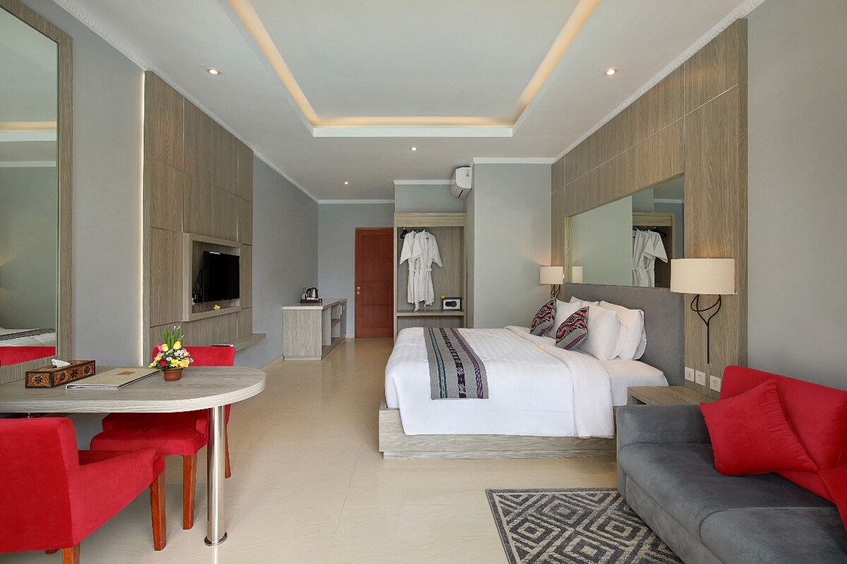 A spacious room features a comfortable king-size bed with decorative pillows. A dining table for two is positioned nearby, accompanied by red chairs. A flat-screen TV is mounted on the wall, and large mirrors enhance the sense of openness. Soft natural light fills the area.