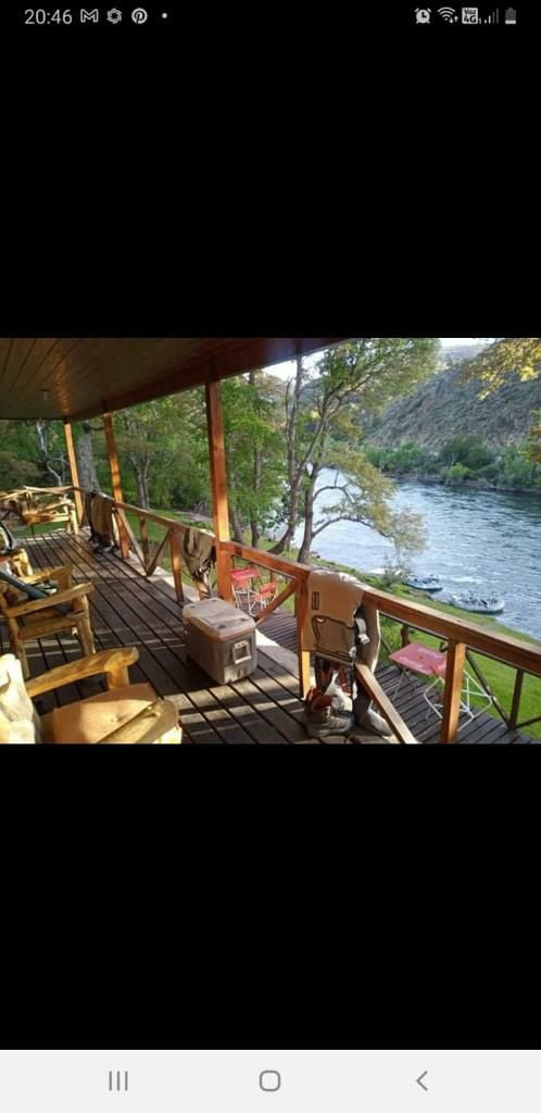 A spacious balcony is shown, featuring wooden chairs arranged along the railing. A cooler is placed beside the railing, and the view reveals the flowing Alumine River below, surrounded by lush greenery and rocky banks.