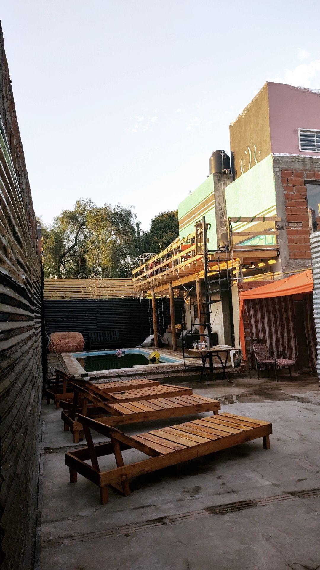 A shared outdoor area features a refreshing pool surrounded by wooden lounge chairs. To one side, vibrant buildings provide a colorful backdrop, while a patio area offers additional seating under a canopy. Access to the space is organized for communal use, serving as an inviting retreat.