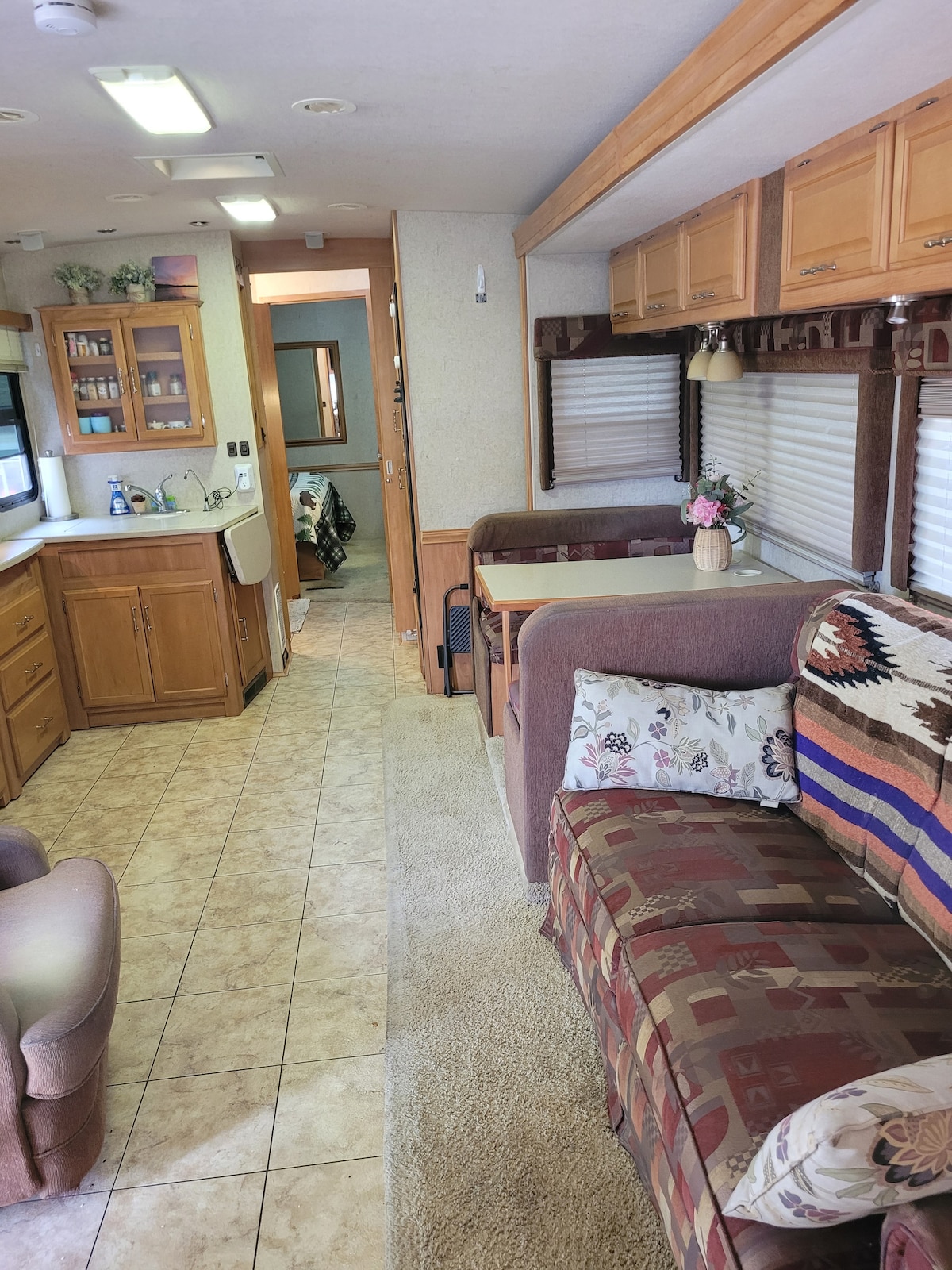 An interior view of the stationary RV showcases a spacious living area with comfortable seating, including a patterned couch and an armchair. A dining area with a table is visible, along with a kitchenette fitted with wooden cabinetry. Light filters through the windows dressed in blinds.