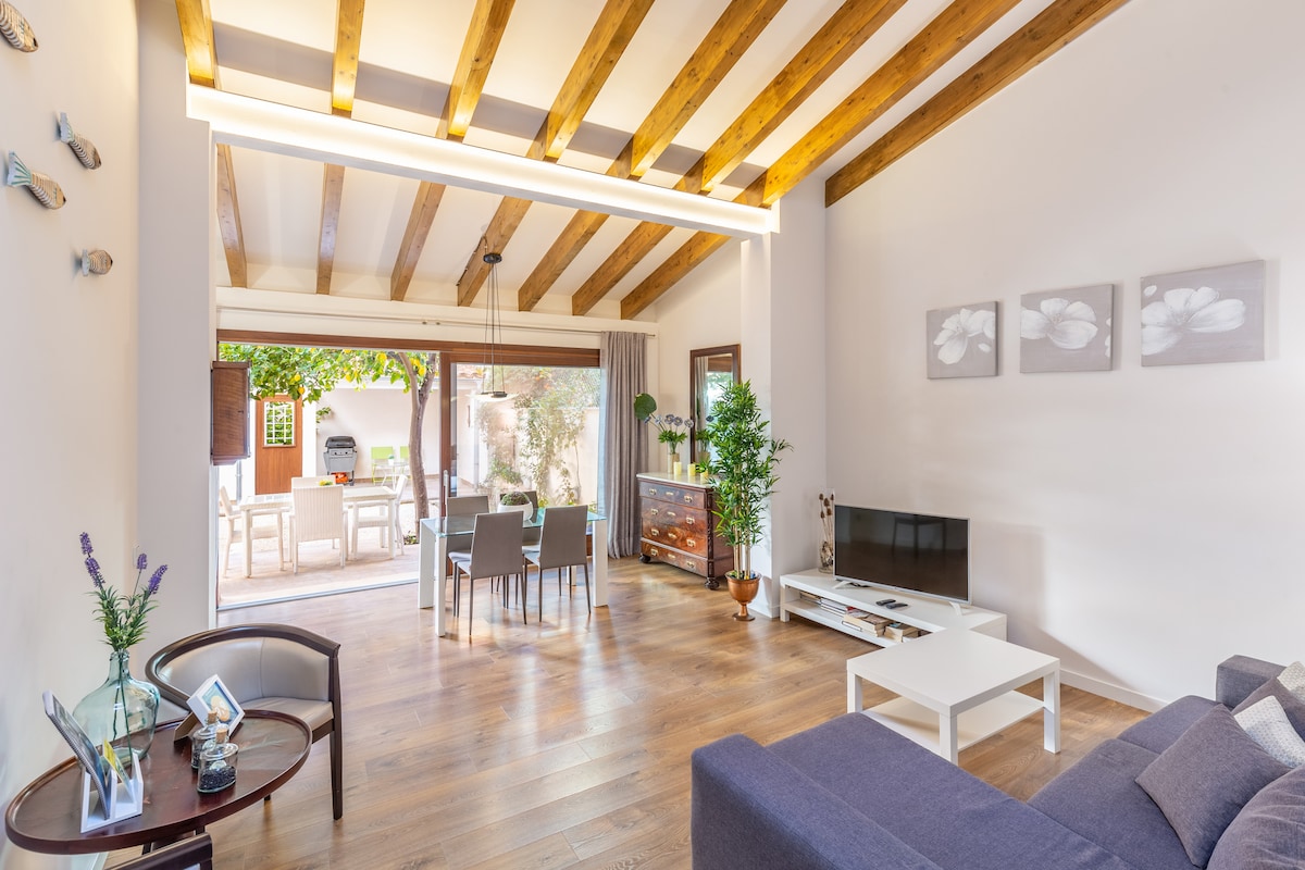 A bright living area features wooden beams overhead, with a sectional sofa and coffee table positioned beside a television. A dining area with six chairs is visible in the background, complemented by a view of the outdoor terrace through sliding glass doors.