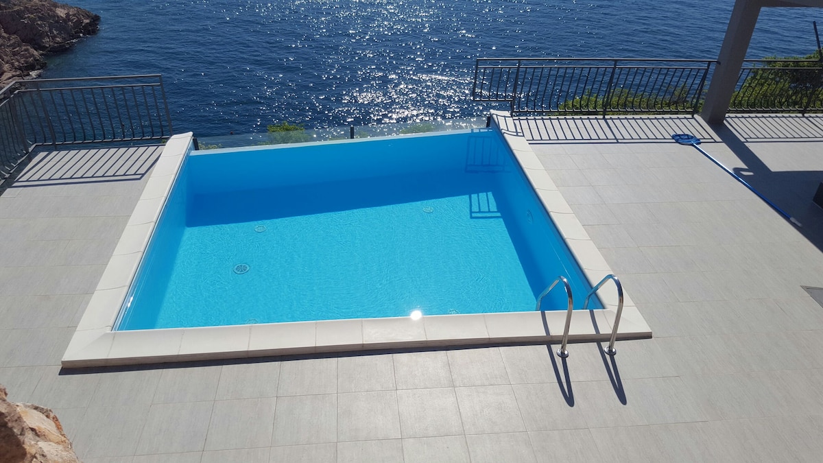 A private swimming pool with clear turquoise water is set against a backdrop of shimmering sea waves. The pool area features a smooth tiled deck and stainless steel steps leading into the water, providing a relaxing space to unwind and enjoy the scenic view.
