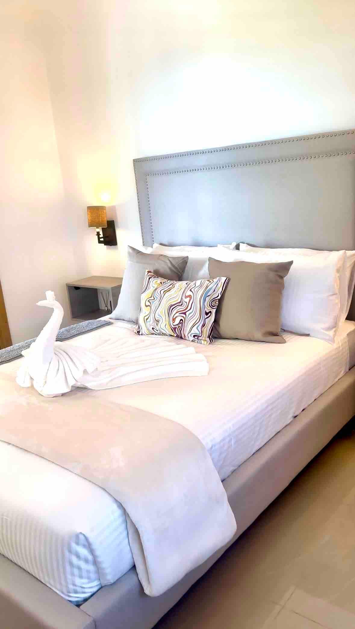 A cozy sleeping area features a spacious bed with soft bedding, accentuated by decorative pillows. A neatly arranged towel sculpture resembling a swan rests on the bed. A bedside lamp provides subtle lighting, adding comfort to the serene ambiance.