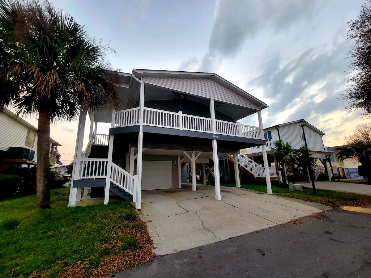 The home is situated on a raised foundation, featuring a wrap-around porch with white railings. Two palm trees frame the entrance, and a concrete driveway leads to a garage area below. The exterior is painted in light hues, with a partially cloudy sky in the background.