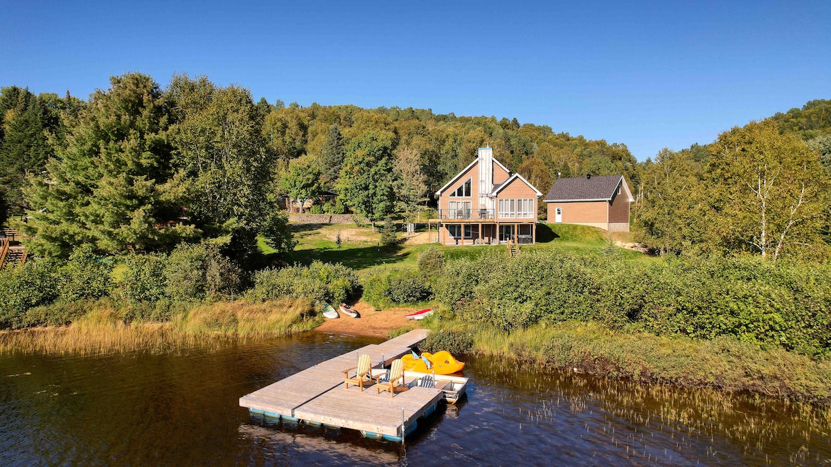 A view of the chalet on the edge of the lake is captured, showcasing its modern design and surrounding nature. A private dock is visible in front, with chairs and kayaks positioned nearby. Lush greenery and a sandy area enhance the scenic lakeside setting.