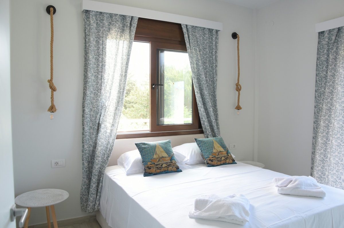 A bright bedroom is revealed, featuring a spacious bed with decorative pillows and fresh towels neatly placed. The window is adorned with patterned curtains that allow natural light to enter, showcasing views of the surrounding greenery.