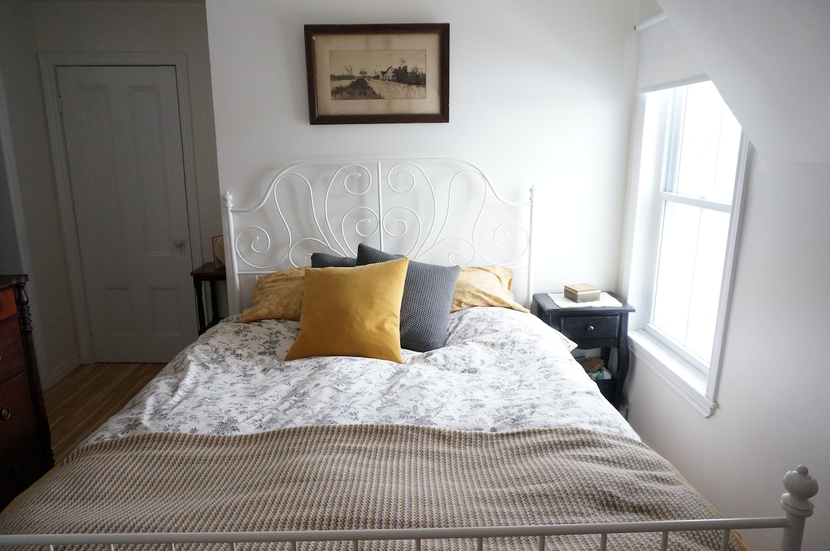 A cozy sleeping area features a white metal bed framed with soft linens and decorative pillows in shades of gray and yellow. A small bedside table holds a few books, and natural light fills the space through the window adorned with a light curtain.