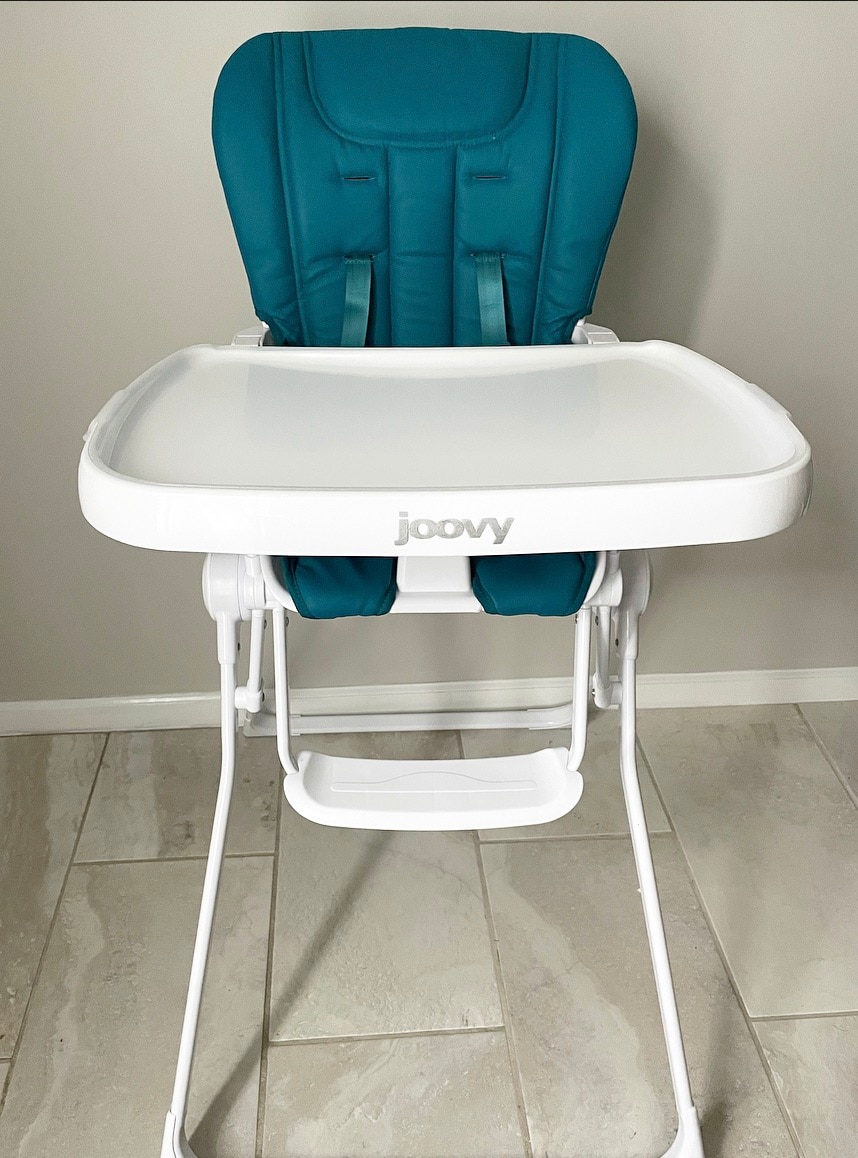 A high chair is displayed in a neutral-toned room, featuring a teal padded seat for comfort and a large, easy-to-clean white tray. The chair stands on a white frame, positioned on a tiled floor, providing a safe and accessible seating option for young children.