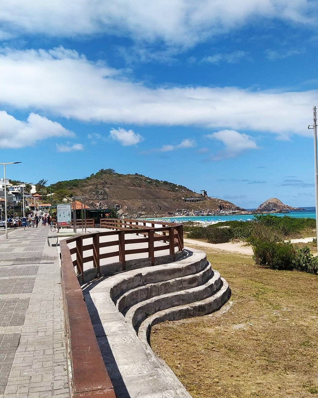 A scenic beachfront view is captured, featuring a walkway lined with wooden railings and gentle, curving seating areas. The coastline stretches into the distance, where green hills meet a clear, blue sky dotted with clouds. Boats are visible in the water, hinting at recreational activities.