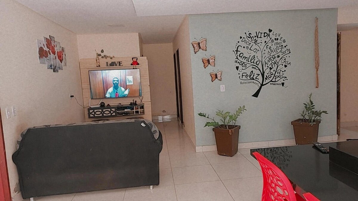 A living area features a black couch positioned near a television, which is displaying content. Wall decor includes a tree design with words representing family and happiness. Two potted plants are situated on the floor, contributing to the space's relaxed atmosphere.