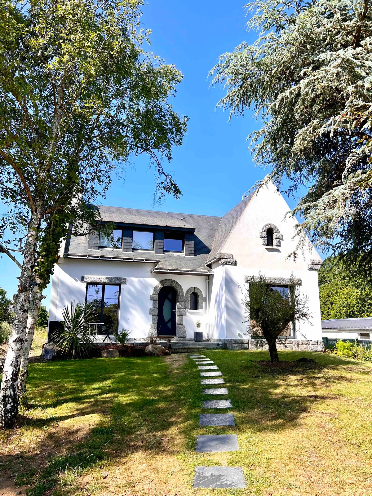 A charming house is set against a clear blue sky, featuring a peaked roof and modern windows. A stone pathway leads from the front yard, surrounded by trees and greenery, to the entrance, highlighting the peaceful outdoor setting.