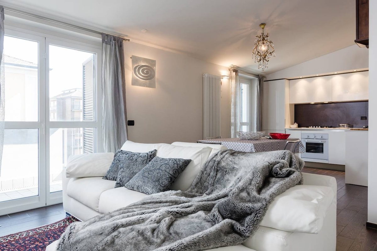 A bright and cozy living area features a white sectional sofa adorned with decorative pillows and a soft, textured throw. Natural light streams in through large windows, illuminating the open kitchen and dining space in the background, which includes modern appliances and minimalistic decor.