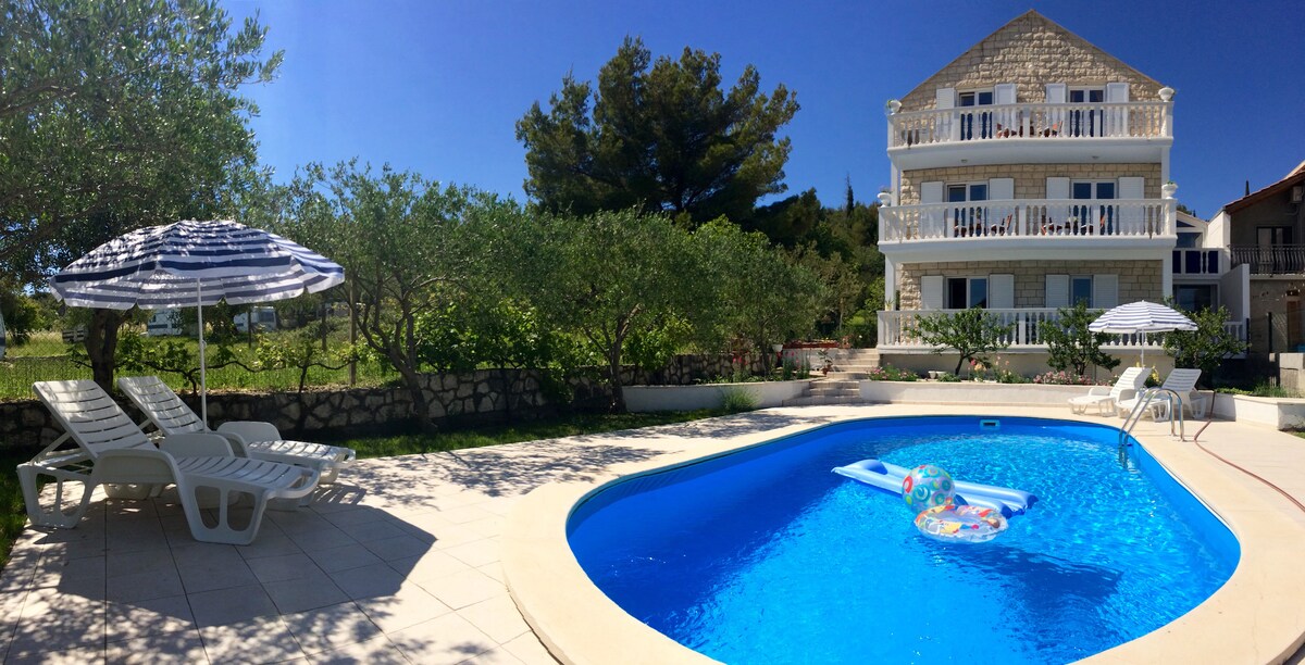 A clear blue pool is surrounded by lounge chairs and umbrellas, set within a well-maintained outdoor area. Lush greenery and trees provide shade in the background, while a three-story building is visible, contributing to the serene atmosphere of the space.