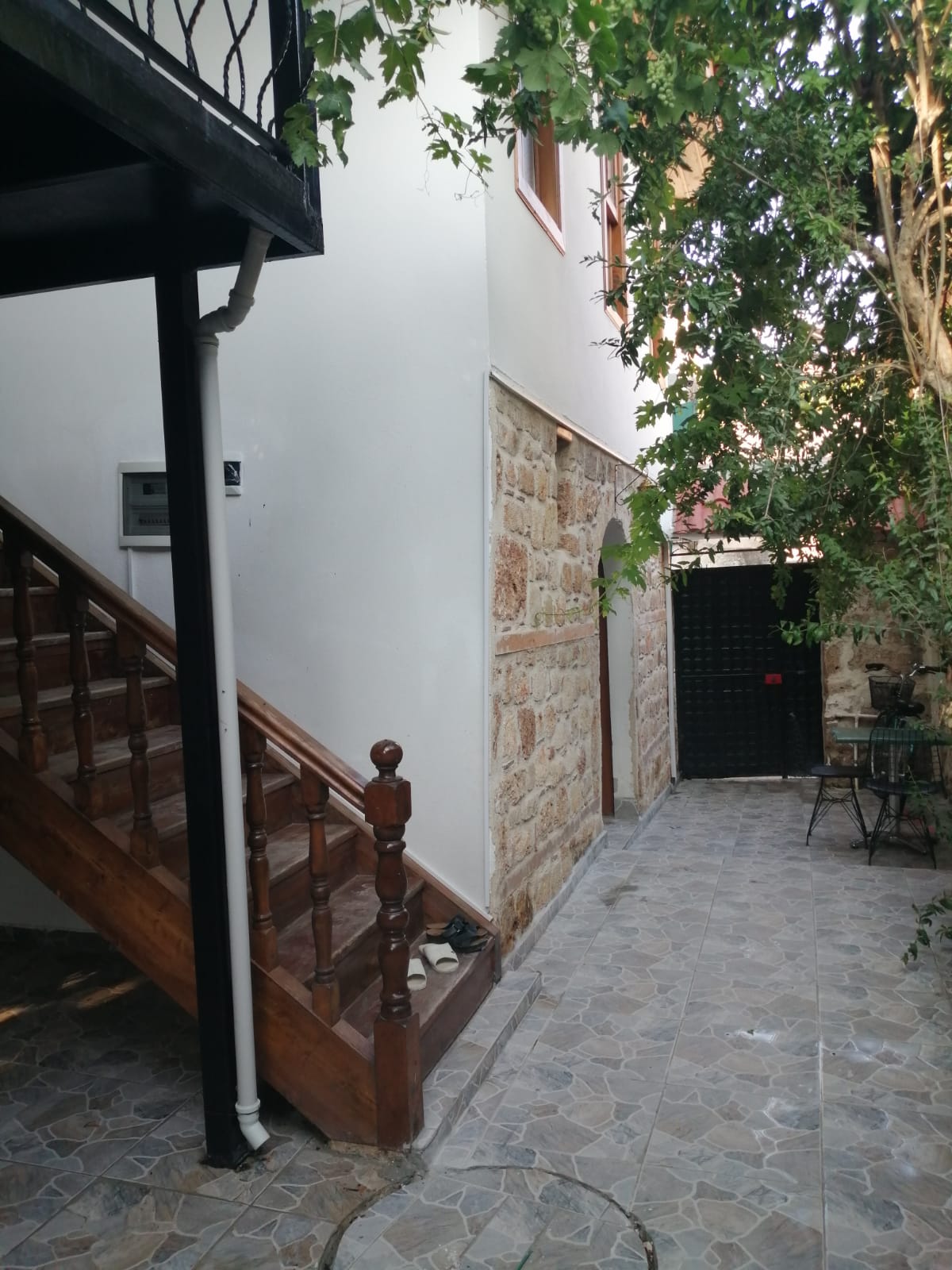 A rustic stone wall complements the wooden staircase leading to an upper level. The ground is covered in patterned tiles, surrounded by lush greenery. A black gate marks the entry to a private area, while a small seating arrangement is visible in the background.