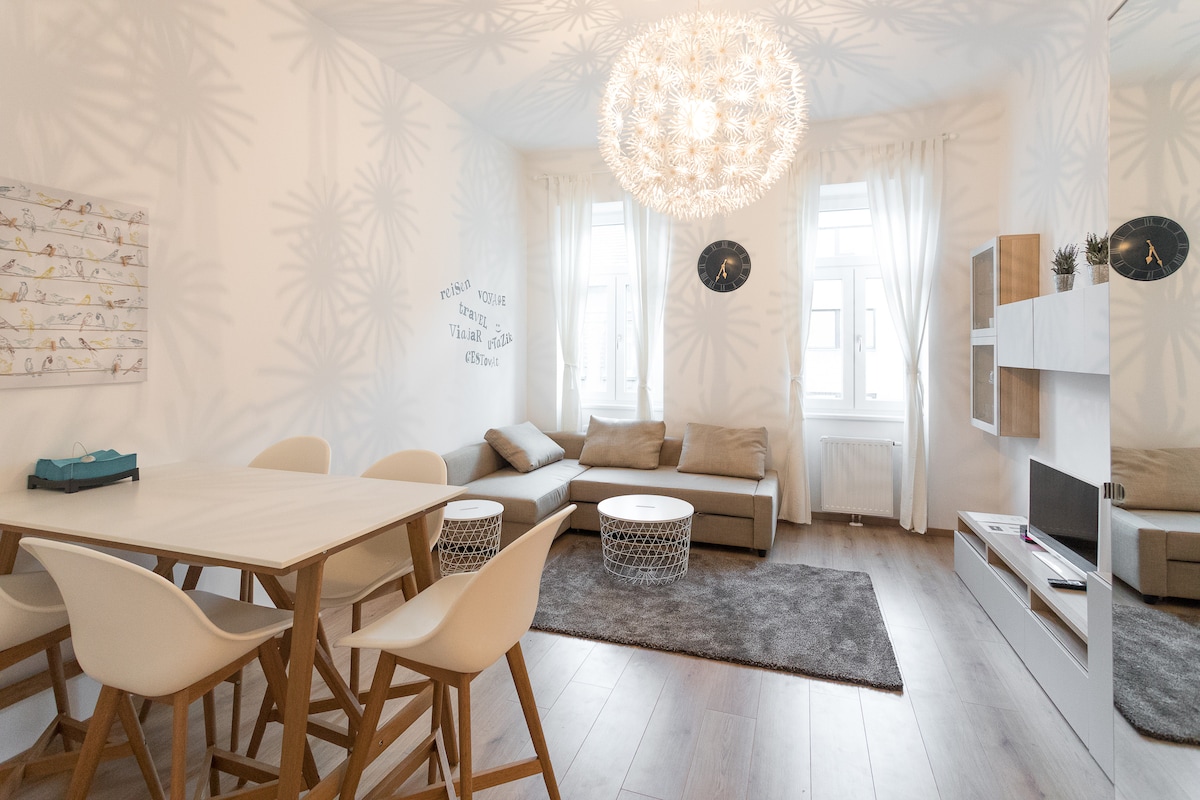 A modern living room features a cozy sectional sofa and a stylish light fixture casting intricate shadows on the walls. A dining table with white chairs is positioned nearby, with two accent tables and a soft area rug completing the space.
