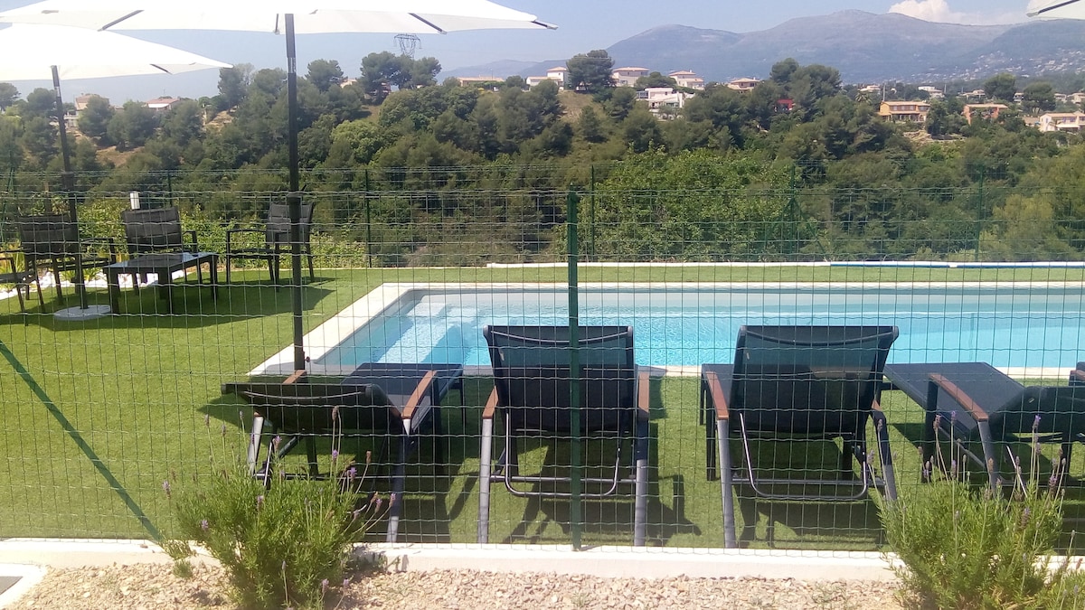 A spacious outdoor area features a swimming pool surrounded by sun loungers and umbrellas. Lush greenery is visible in the background, providing a scenic view of the landscape. A secure fence ensures privacy and safety for guests enjoying the outdoor amenities.