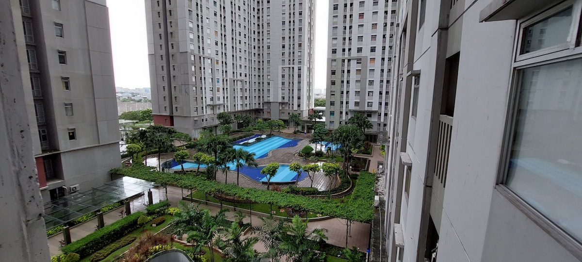 A serene outdoor area is visible, featuring two swimming pools surrounded by neatly arranged greenery and palm trees. Tall residential buildings frame the space, providing a sense of tranquility amidst the urban setting.
